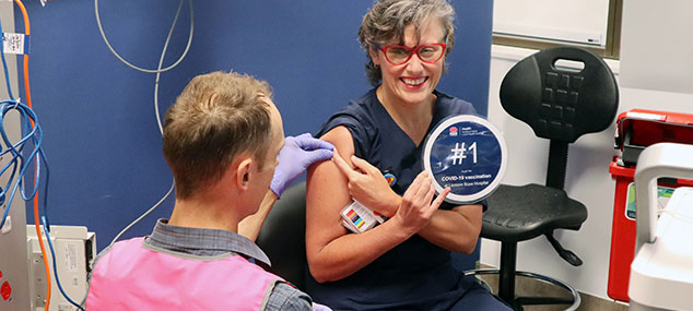 Registered Nurse, Rachel holds a sign saying '1st COVID vaccination' at Lismore Base Hospital.